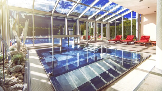 Una piscina interior con paredes de vidrio transparente, sillas de descanso rojas y vistas al exterior.