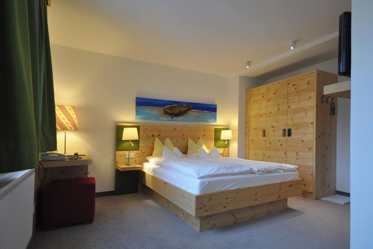 A wooden bed in a hotel room with a painting of a boat on the wall, two bedside tables with lamps, and a wooden wardrobe.