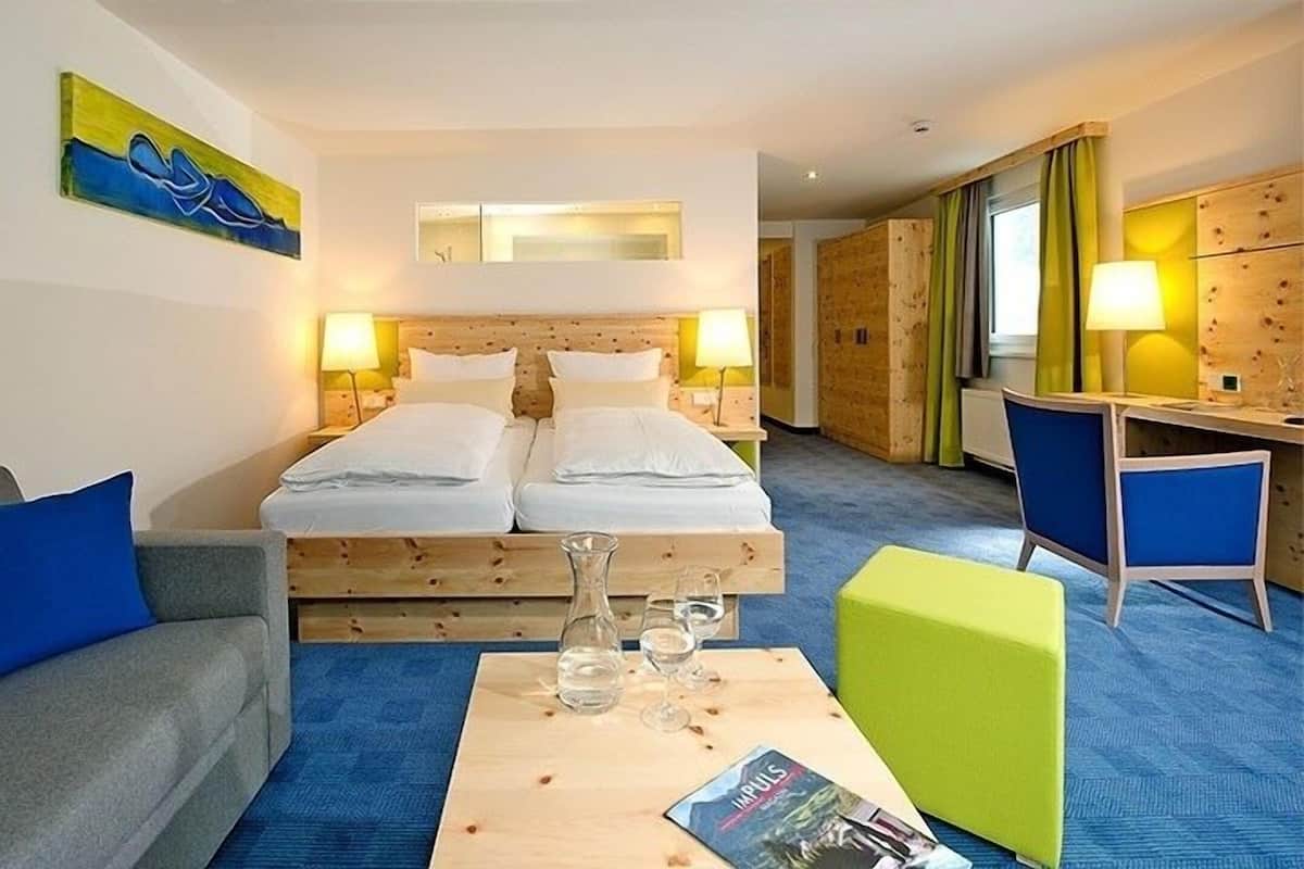 A hotel room with a double bed, a desk, a chair, a coffee table with a magazine, and a painting on the wall.