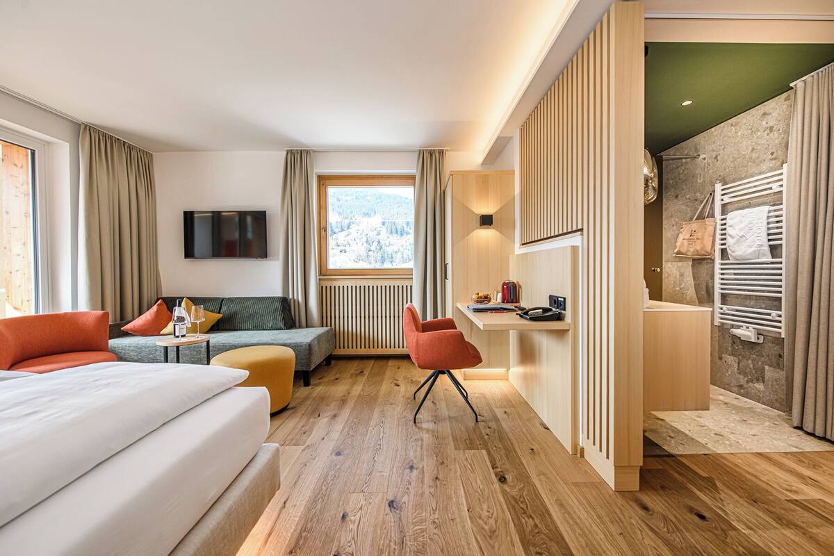 A hotel room with a bed, sofa, chair, TV, window, and a kitchenette area.