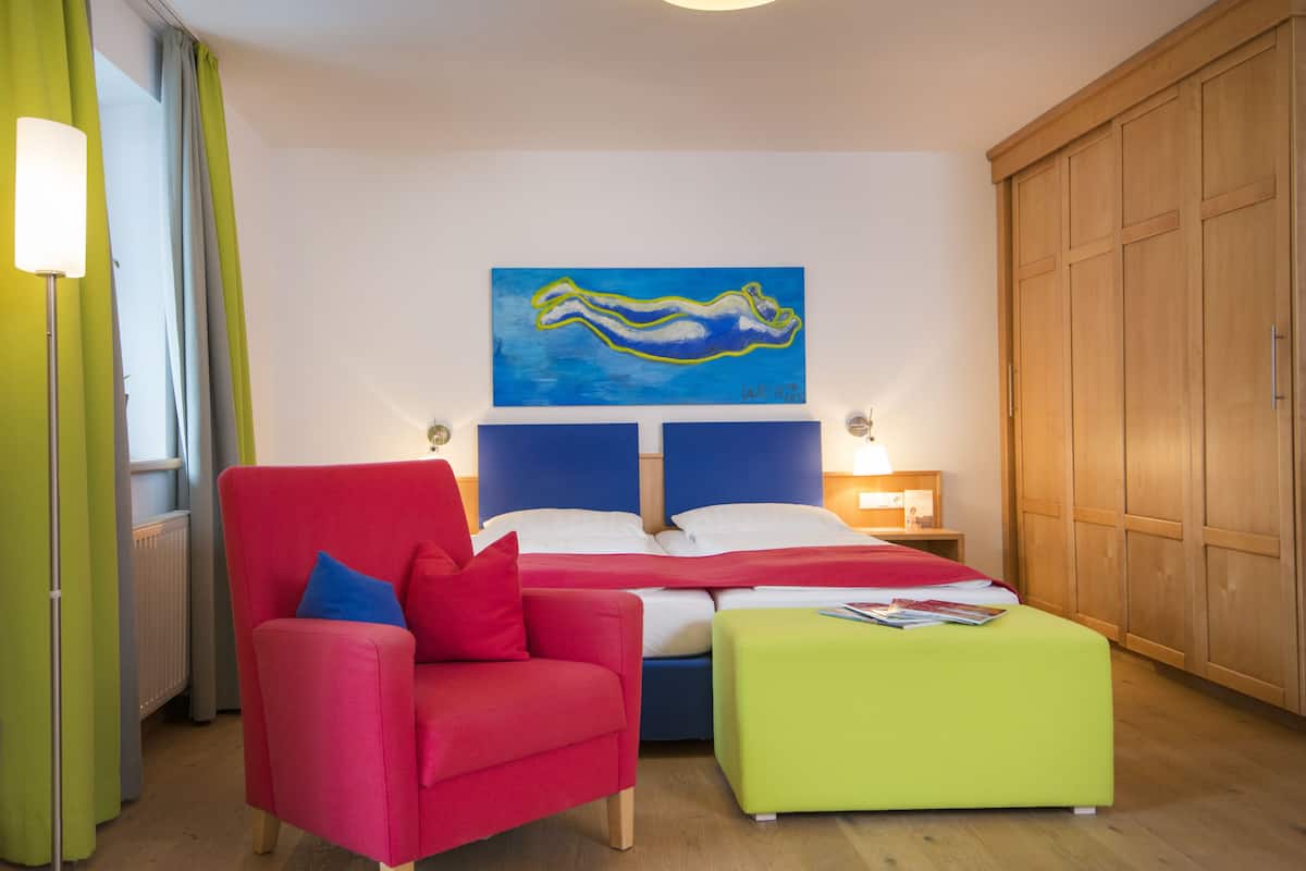 A bedroom with a bed, a red chair, a green ottoman, a painting on the wall, and wooden wardrobe.