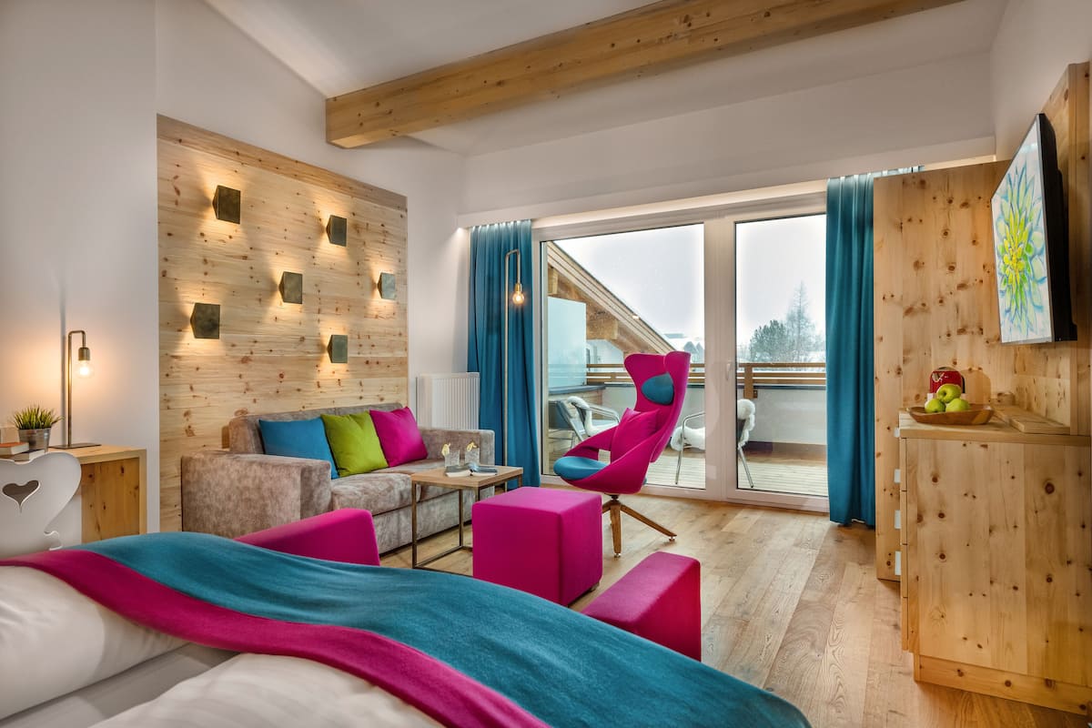 A modern hotel room with a wooden accent wall, a balcony with a view, a sofa, a chair, and a bed with a colorful blanket.