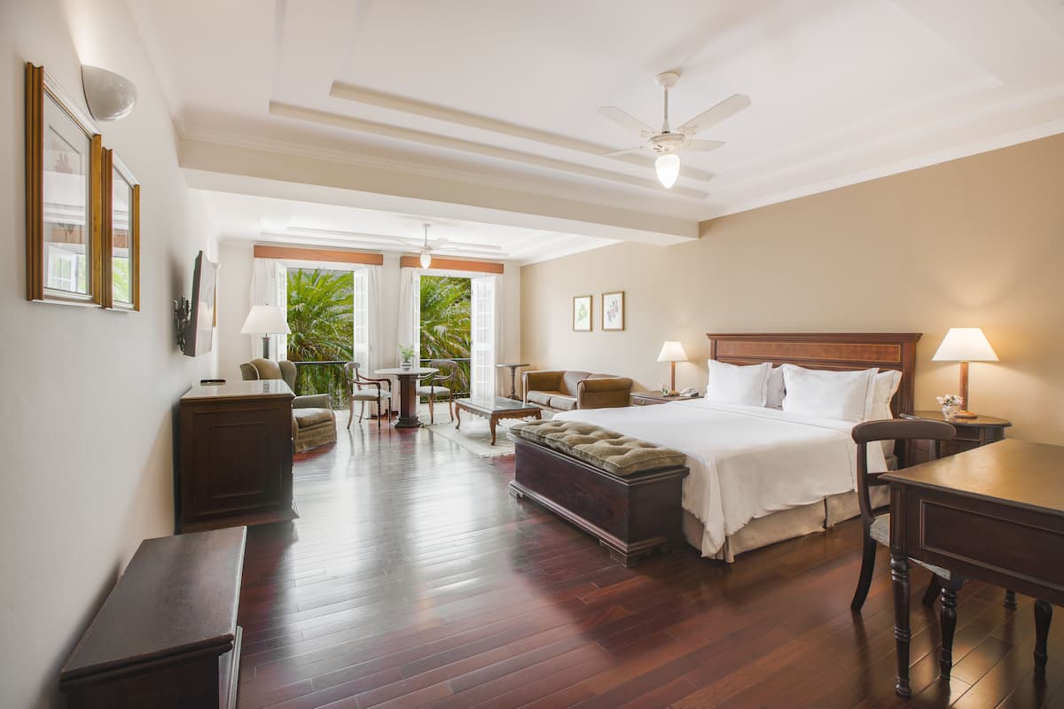 Habitación de hotel con una cama grande, un escritorio, una silla, un sofá y una mesa de comedor. Hay ventanas con cortinas y vistas a zonas verdes.