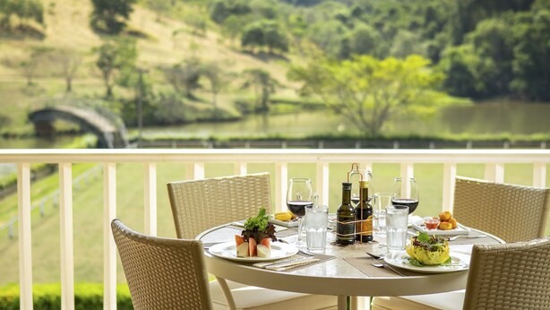 Una mesa de comedor con sillas, una botella de vino, copas y un plato de comida en un balcón con vistas a un paisaje pintoresco con un puente y árboles.
