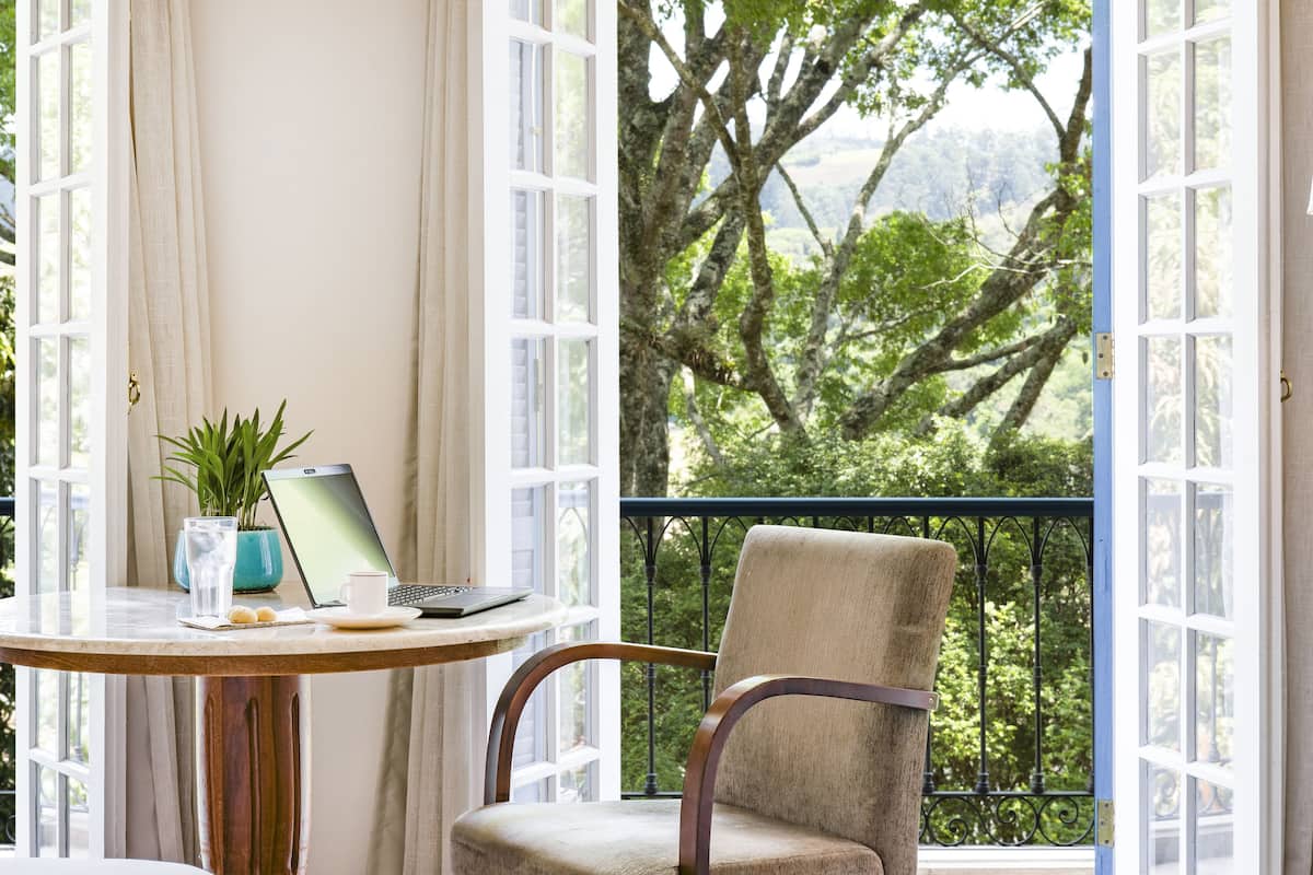 Habitación con vistas a un paisaje verde y exuberante a través de amplios ventanales. Hay una mesa redonda de madera con una computadora portátil, una taza y una planta. Una silla está colocada frente a la mesa.