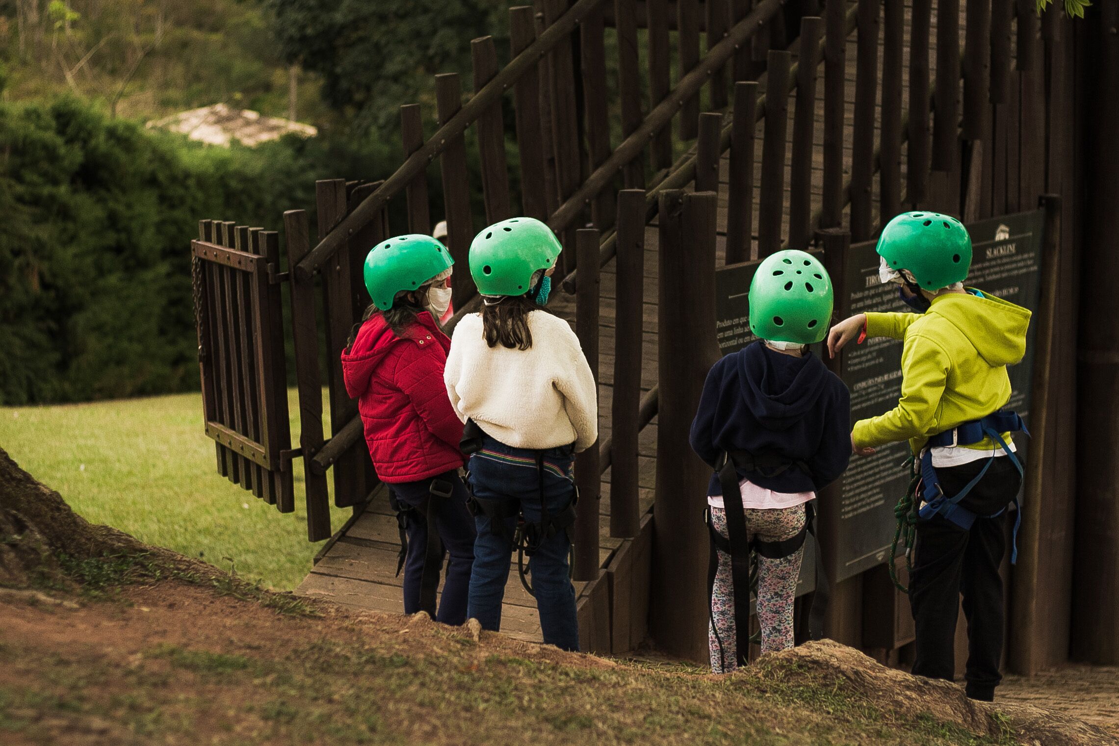 A group of people wearing helmets and harnesses, standing on a wooden bridge with a wooden fence in the background.