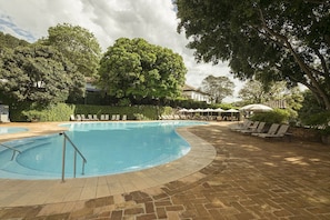 Una piscina con sillones de descanso y sombrillas, rodeada de árboles y un edificio al fondo.