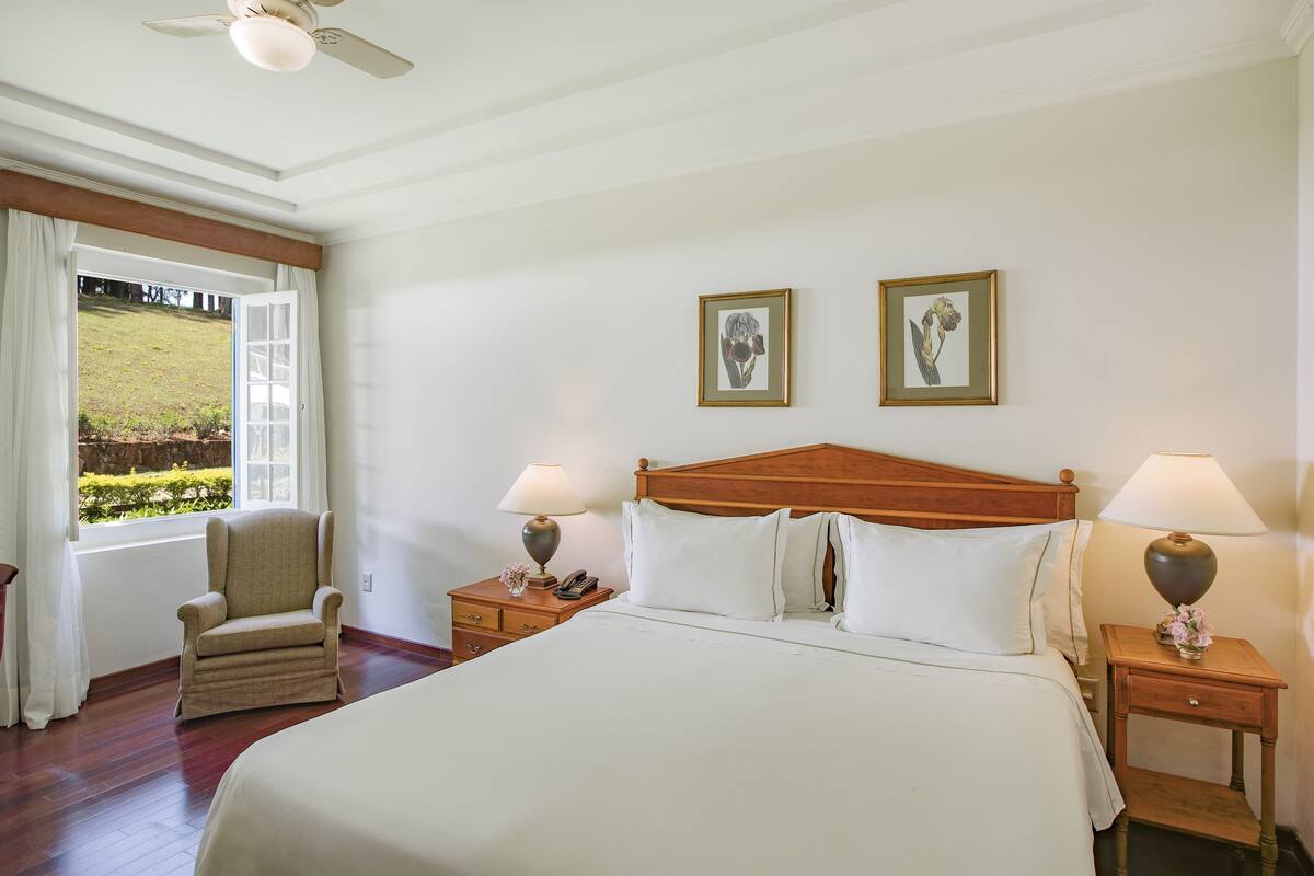 Habitación de hotel con una cama grande, dos mesitas de noche con lámparas, una silla y una ventana con vistas a un jardín.