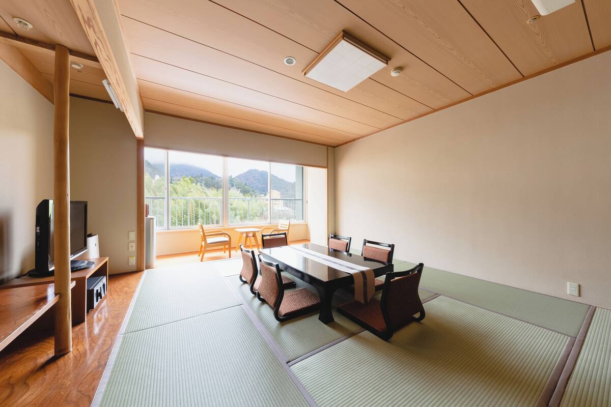 A traditional Japanese-style room with tatami flooring, a low wooden ceiling, and a large window offering a view of mountains.