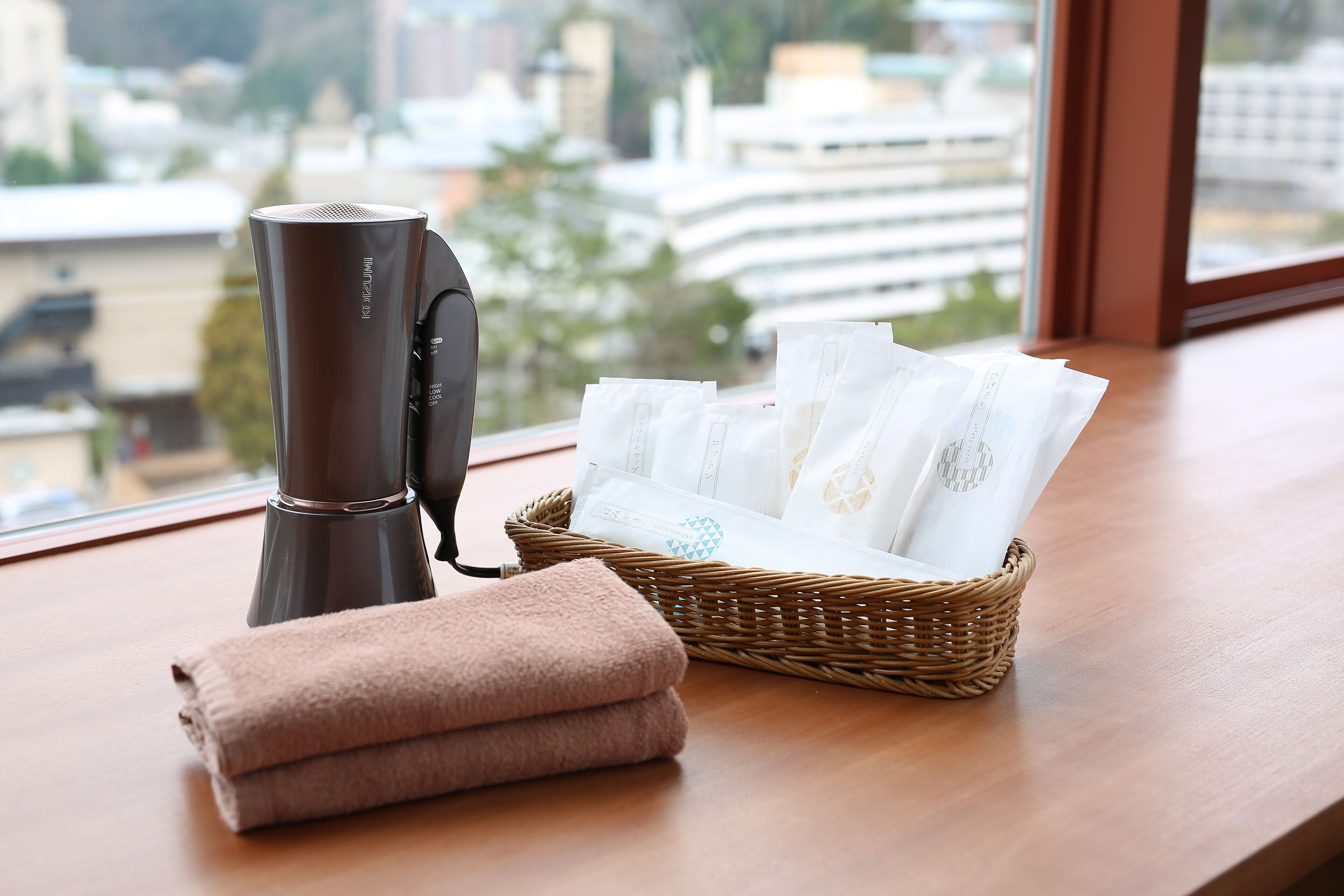A room with a hair dryer, a basket of towels, and a window with a view of buildings.