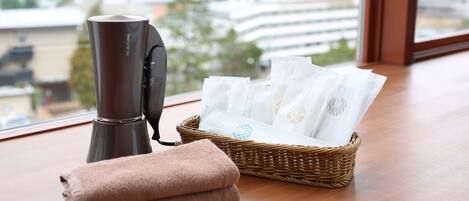 A room with a hair dryer, a basket of towels, and a window with a view of buildings.
