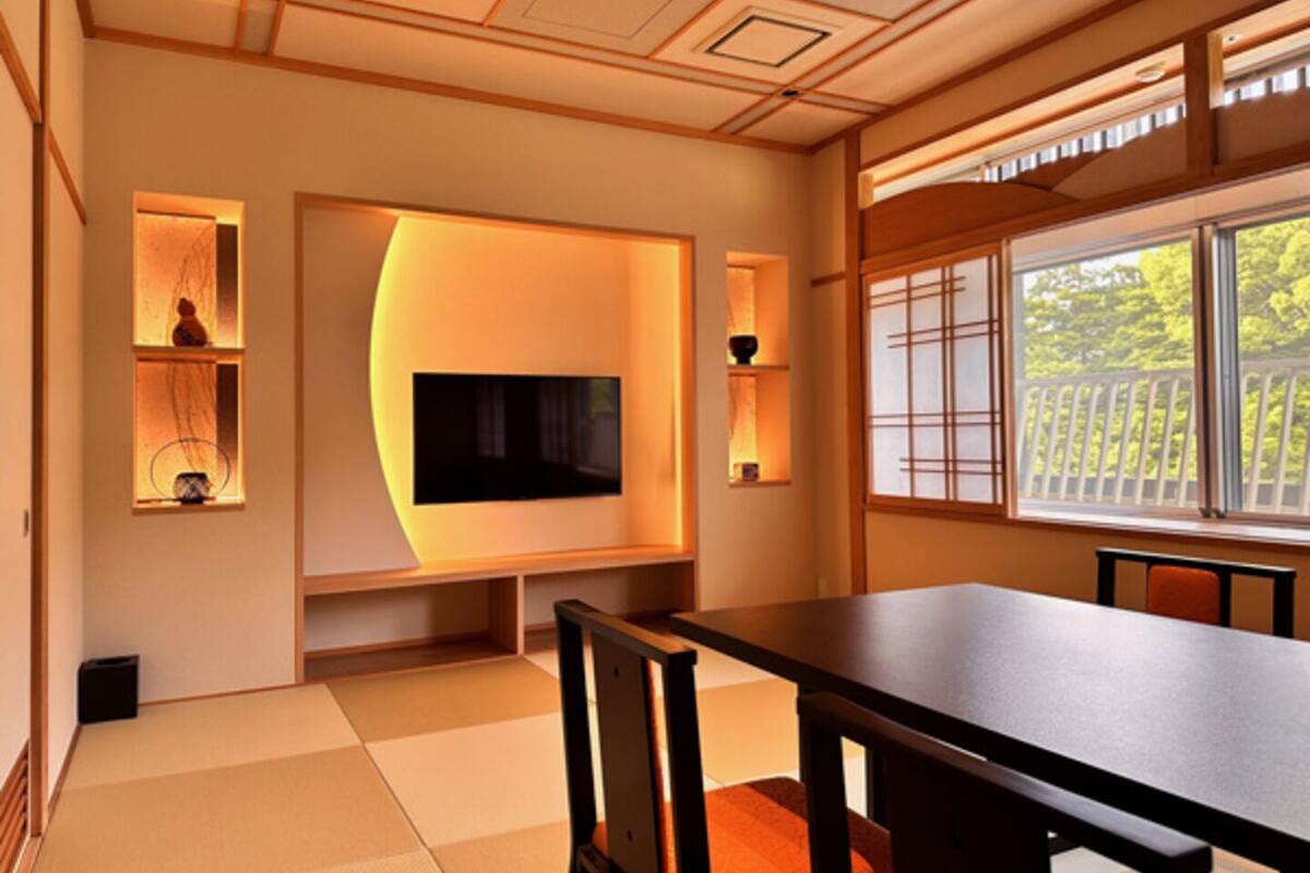 A traditional Japanese-style room with tatami flooring, a low table, chairs, a television, and a window with a view of greenery.