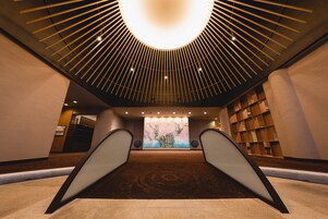 Un moderno vestíbulo de hotel con una escalera central, una gran obra de arte en la pared de fondo y un plafón con una lámpara radial.