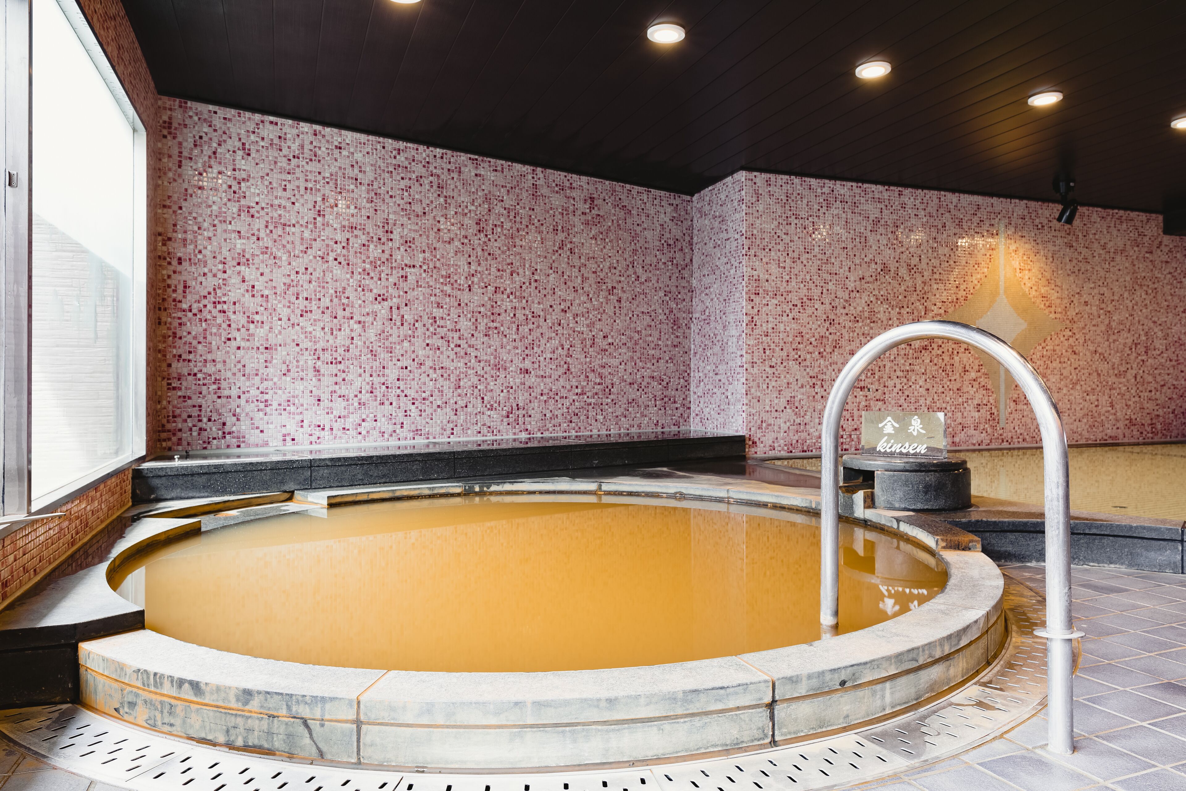 A hot spring bath with a tiled floor, a stone wall with a red mosaic pattern, and a metal faucet.