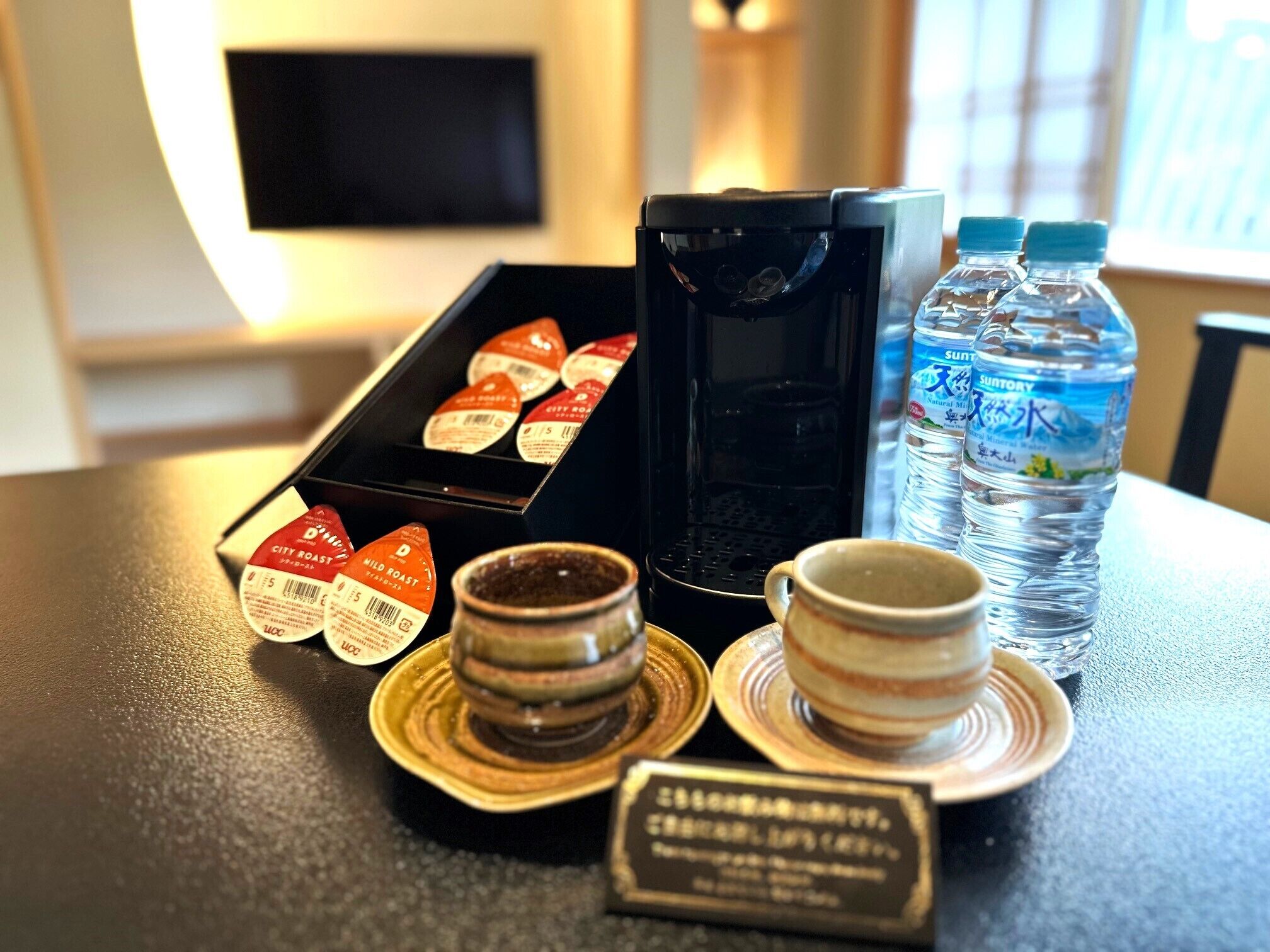A table with a coffee maker, a box of coffee pods, two water bottles, a cup, and a small sign.