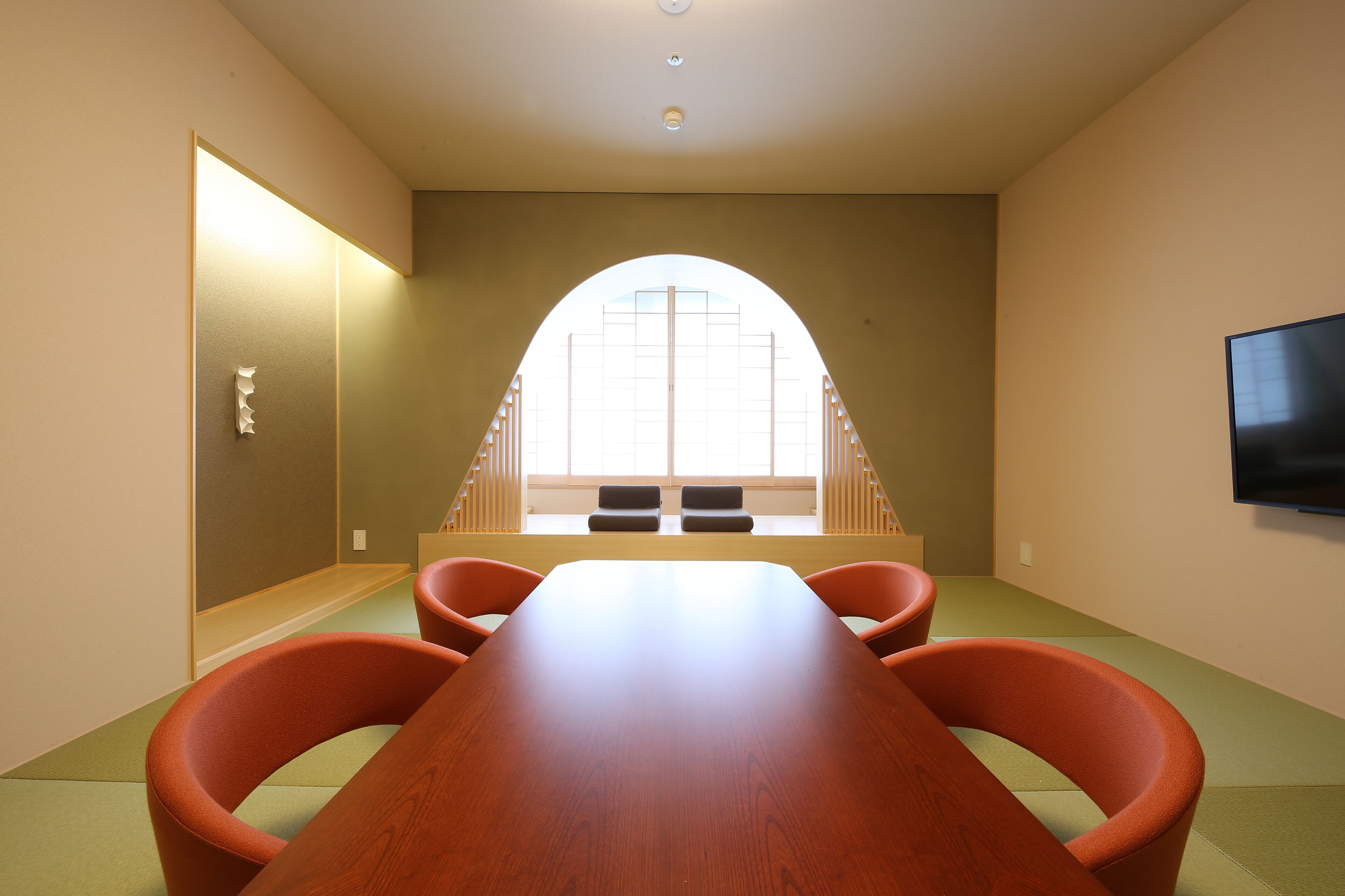 Une salle de conférence avec une longue table, quatre chaises orange, un téléviseur fixé au mur et une grande fenêtre en arc de cercle.