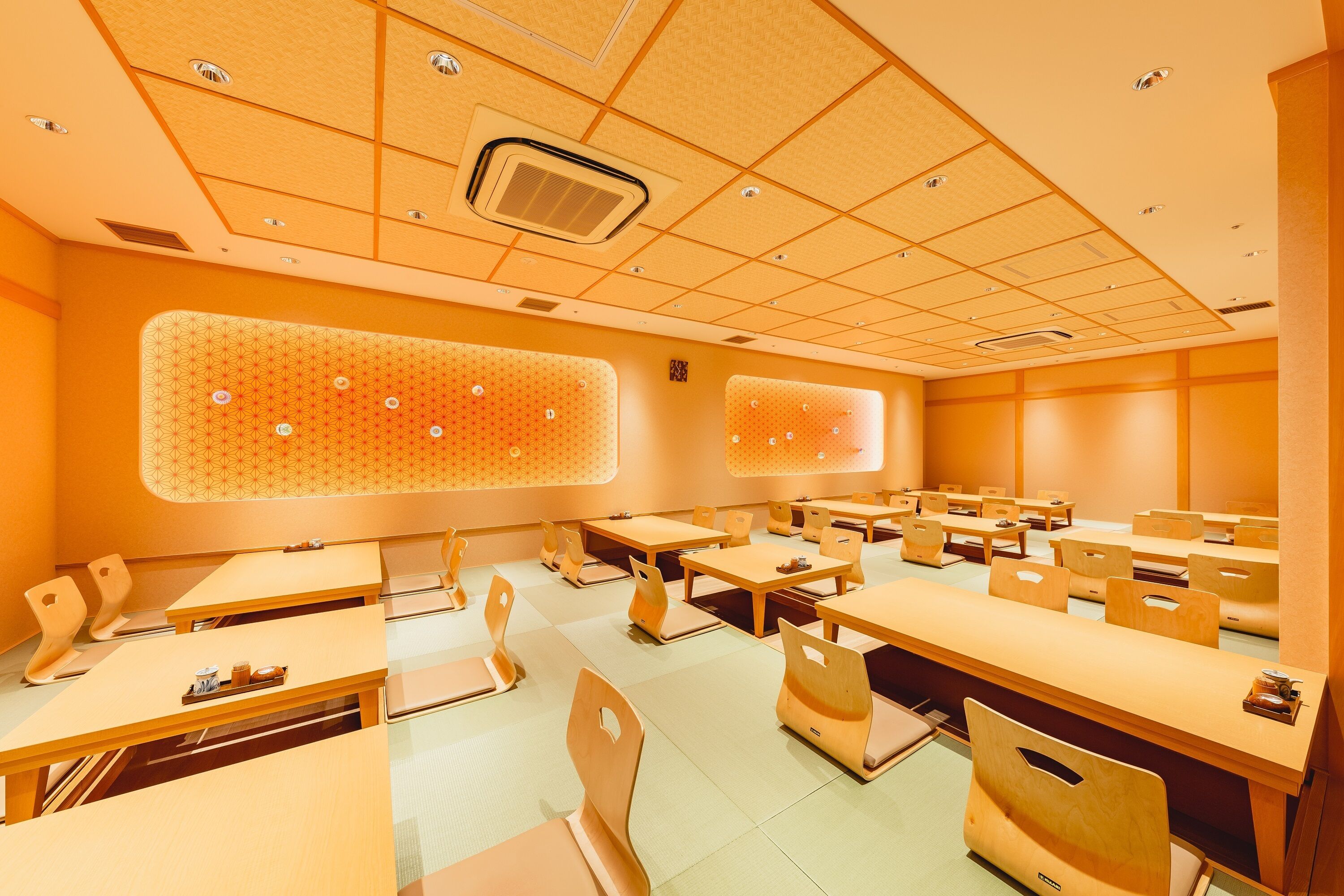 A modern classroom with rows of wooden tables and chairs, a patterned wall, and ceiling lights.