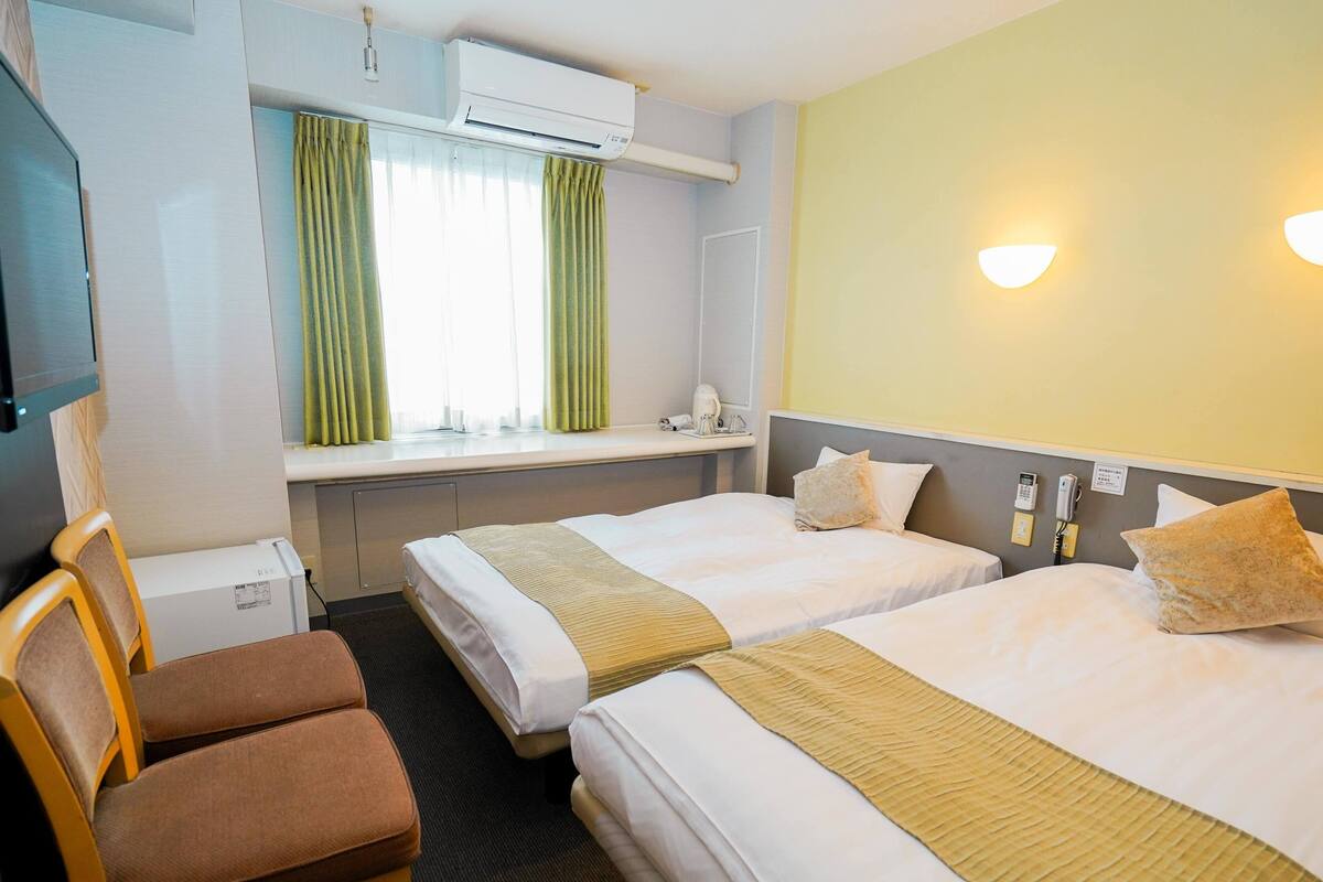 A hotel room with two beds, a desk, a chair, an air conditioning unit, and a window with curtains.