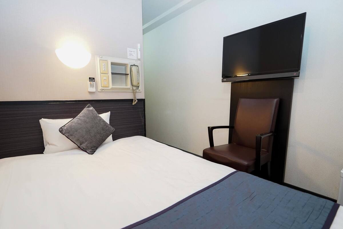A hotel room with a bed, a chair, a TV mounted on the wall, and a telephone.