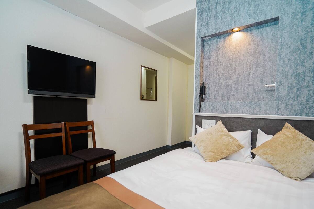 A hotel room with a bed, a TV mounted on the wall, a chair, and a bedside table.