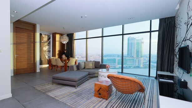 A modern living room with a sofa, ottoman, and coffee table, a dining area with chairs, and a large window with a city view. - Ramee Grand Hotel & Spa (Manama)