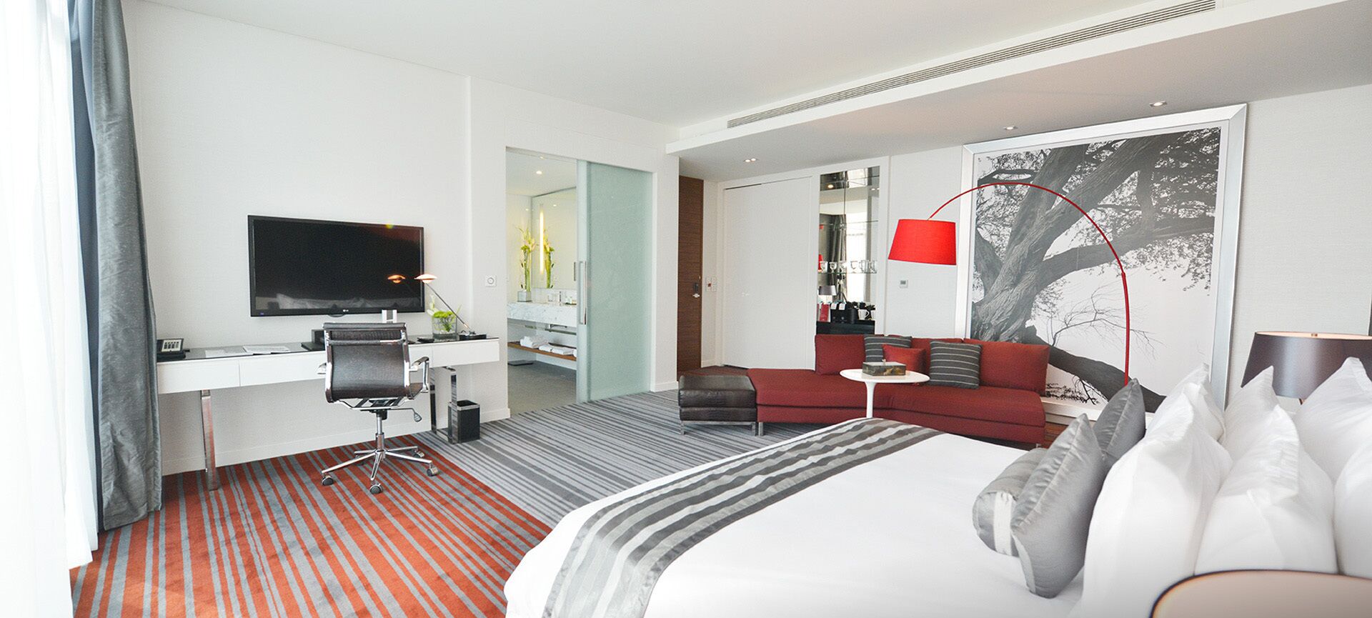 A modern hotel room with a large bed, a desk with a chair, a television, a red sofa, and a red floor lamp.