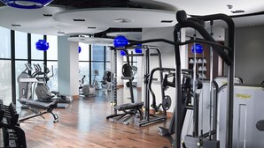 A modern gym with cardio machines, weightlifting equipment, and large windows offering city views. - Ramee Grand Hotel & Spa (Manama)