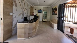 Una zona de recepció amb un taulell de recepció corbat, una zona d'estar amb una banca, una televisió penjada a la paret i un frigorífic.