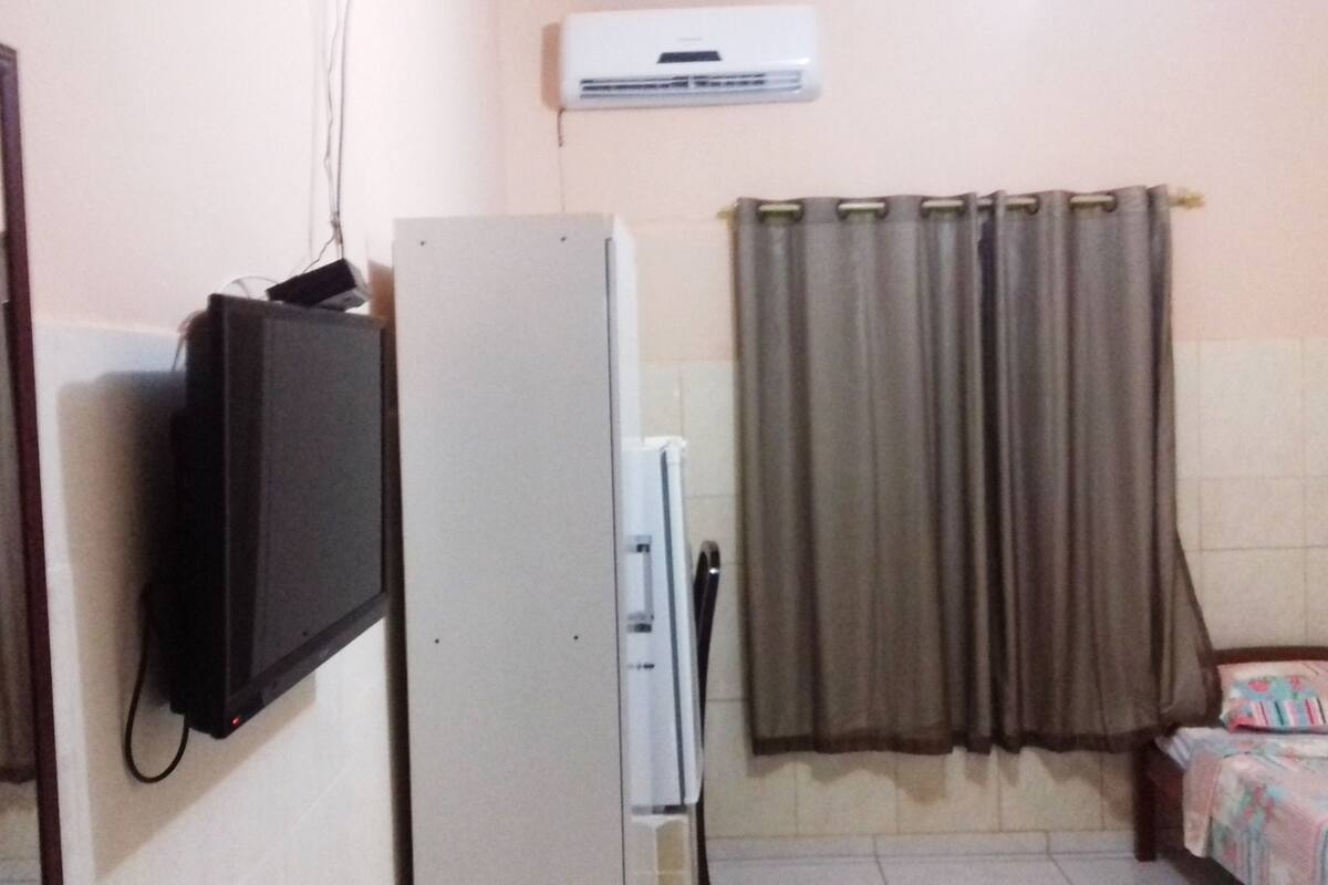 A small room with a refrigerator, a television mounted on the wall, a bed with a floral bedspread, and a window with curtains.