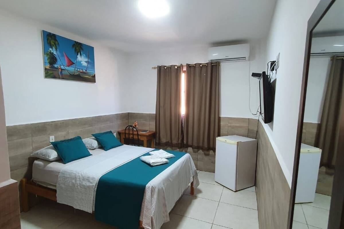 A bedroom with a bed, a small table, a chair, an air conditioning unit, and a painting on the wall.