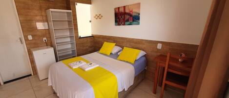 A hotel room with a bed, a wardrobe, a small desk, a chair, a painting, and a wall-mounted shelf.