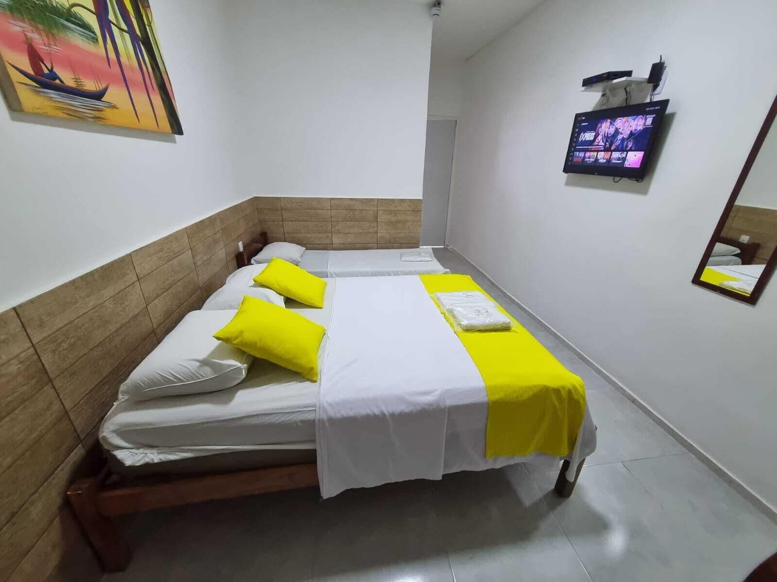A hotel room with two beds, each with white bedding and yellow pillows. There is a flat-screen TV mounted on the wall, and a painting above the left bed.