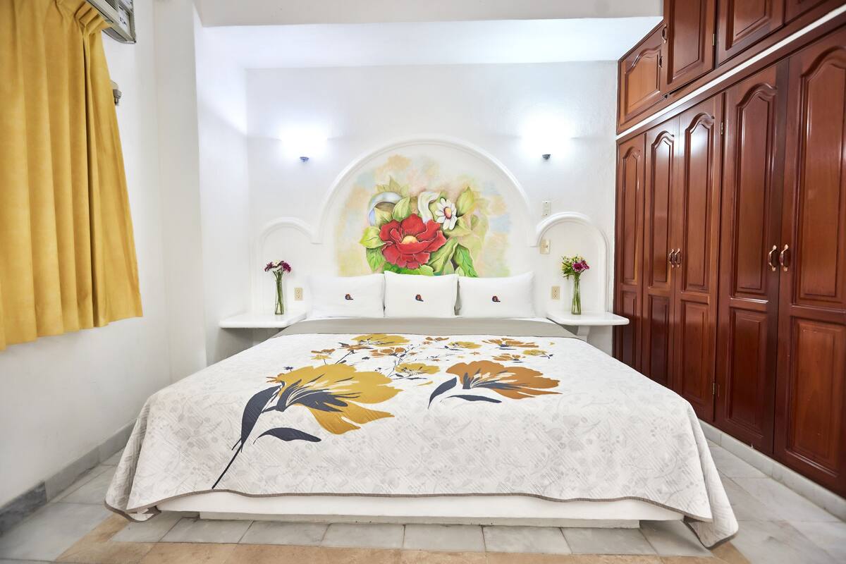 Ein Schlafzimmer mit einem Bett, einem Holzschrank, einem an der Wand befestigten Regal mit Dekoration und einem floralen Gemälde über dem Bett.