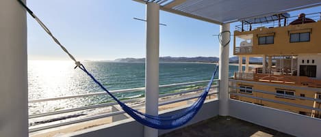 A balcony with a view of the sea, a hammock, and a building with multiple windows.