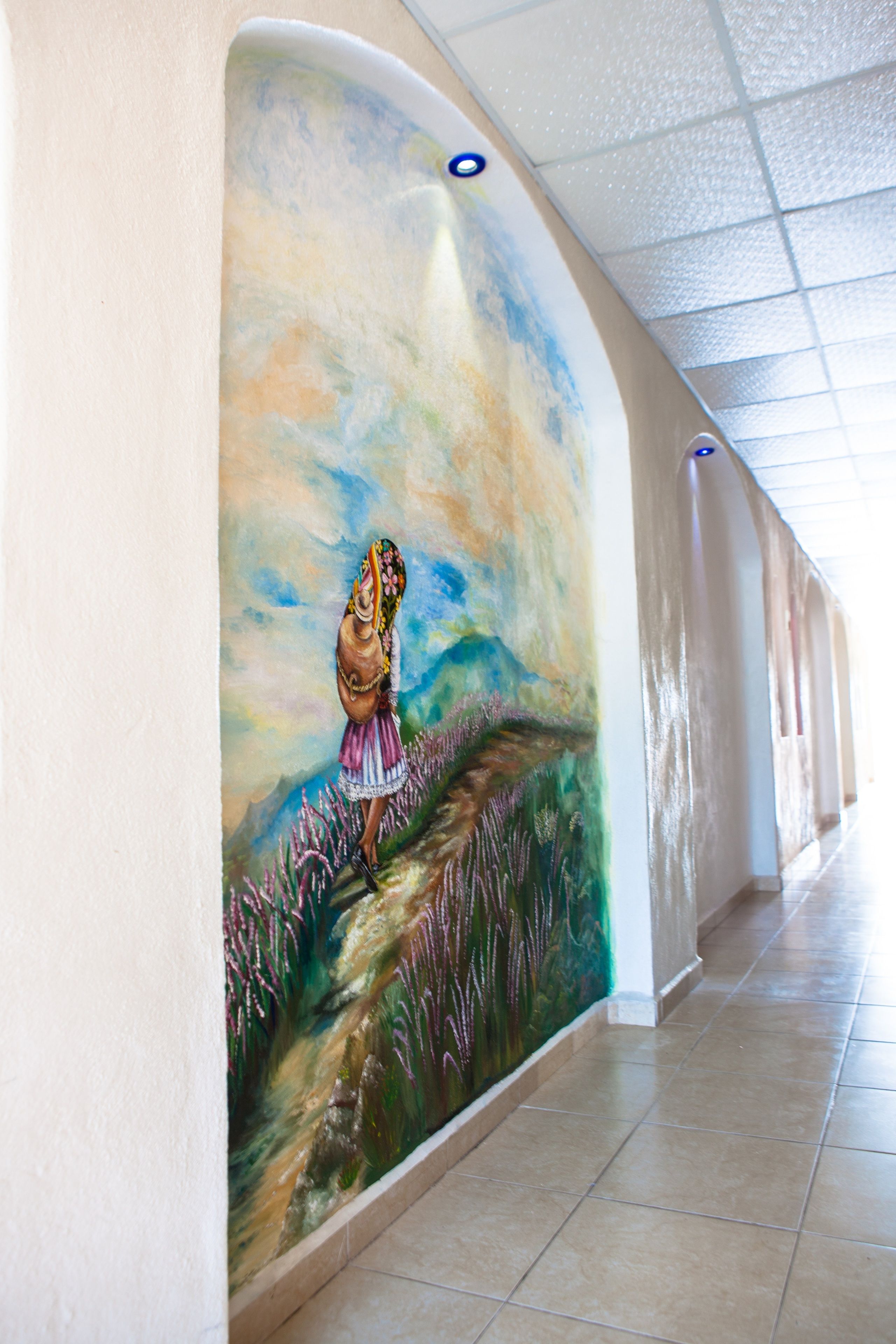 A corridor with a mural of a woman in a field, a tiled floor, and a ceiling with a grid pattern.