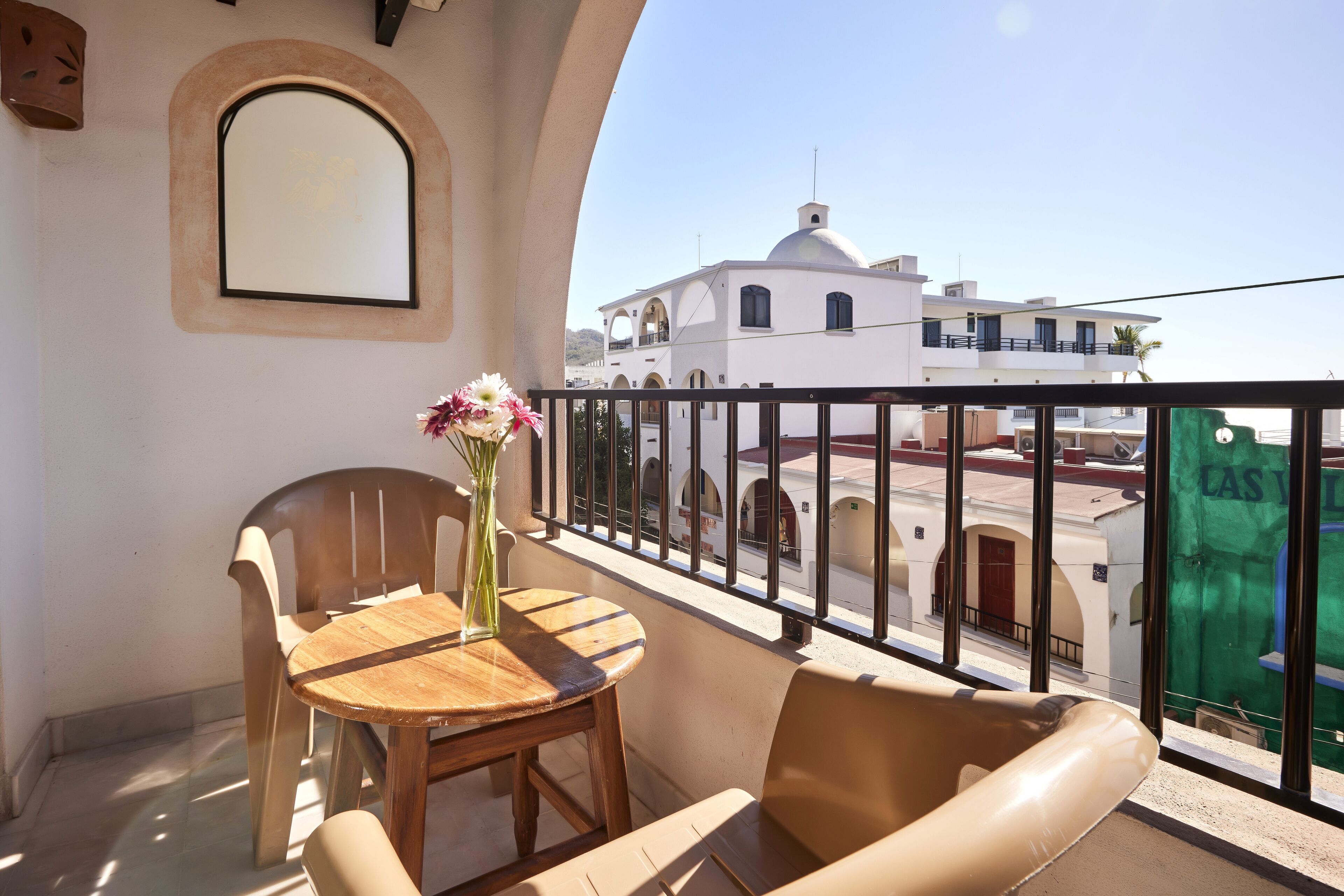 A balcony with a wooden table and chairs, a vase with flowers, and a view of a white building with arches and a dome.