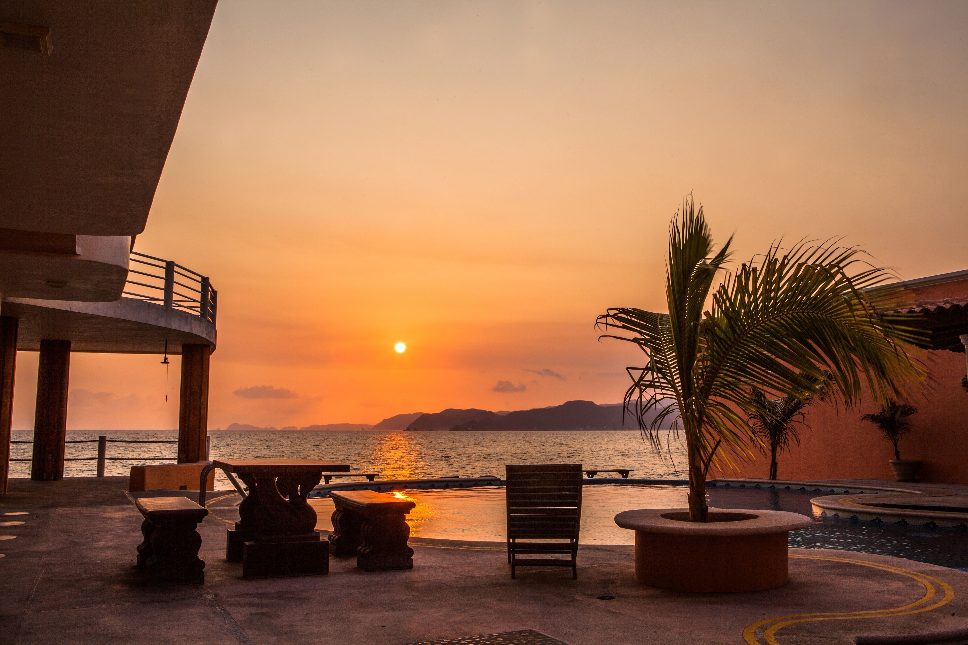 A sunset view from a terrace with a pool, seating, and a palm tree.
