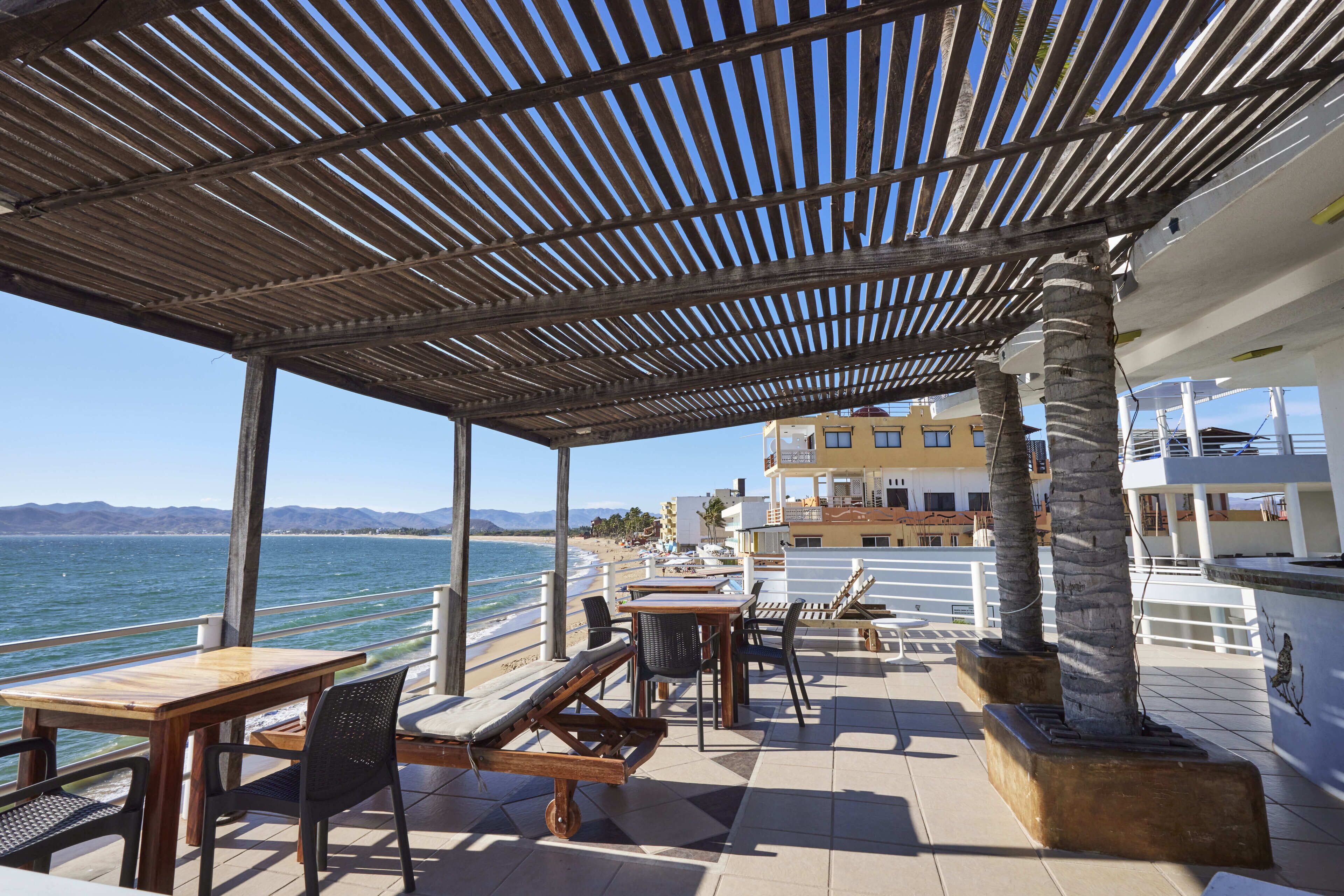 Een overdekt terras met tafels en stoelen, met uitzicht op een strand en de oceaan.