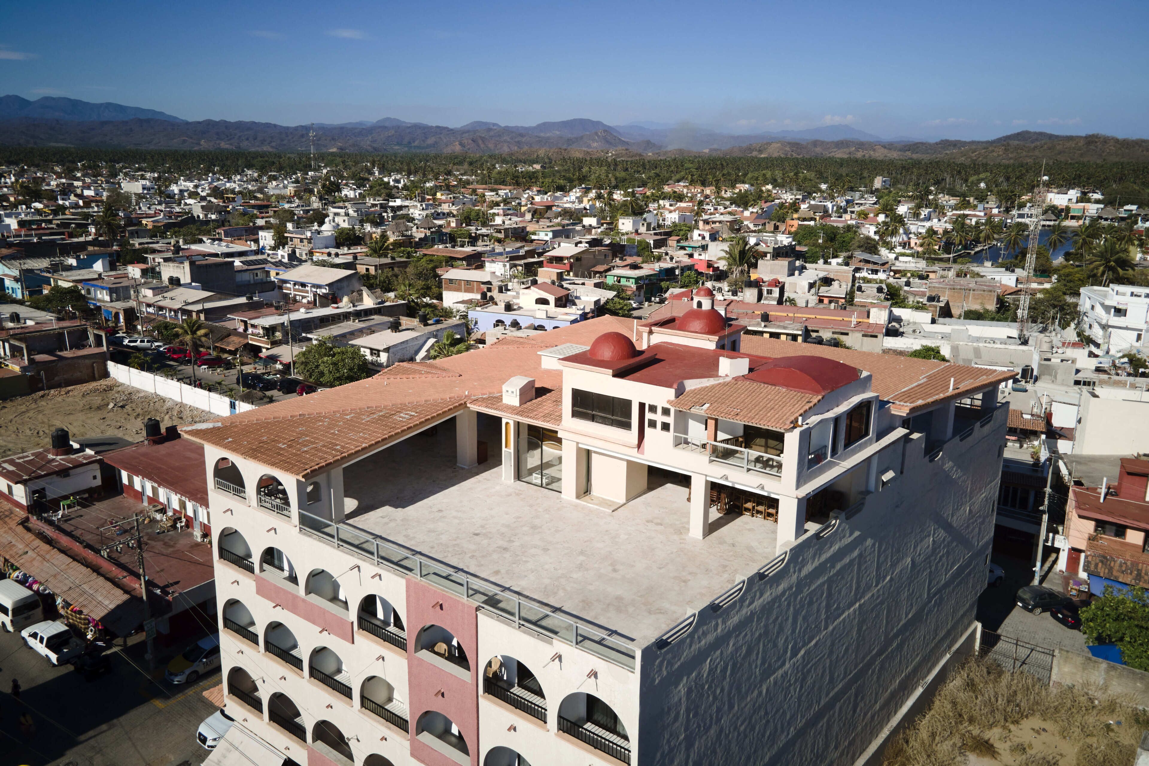 Un edificio de varios pisos con un patio central, rodeado por una zona residencial con casas visibles y montañas al fondo.
