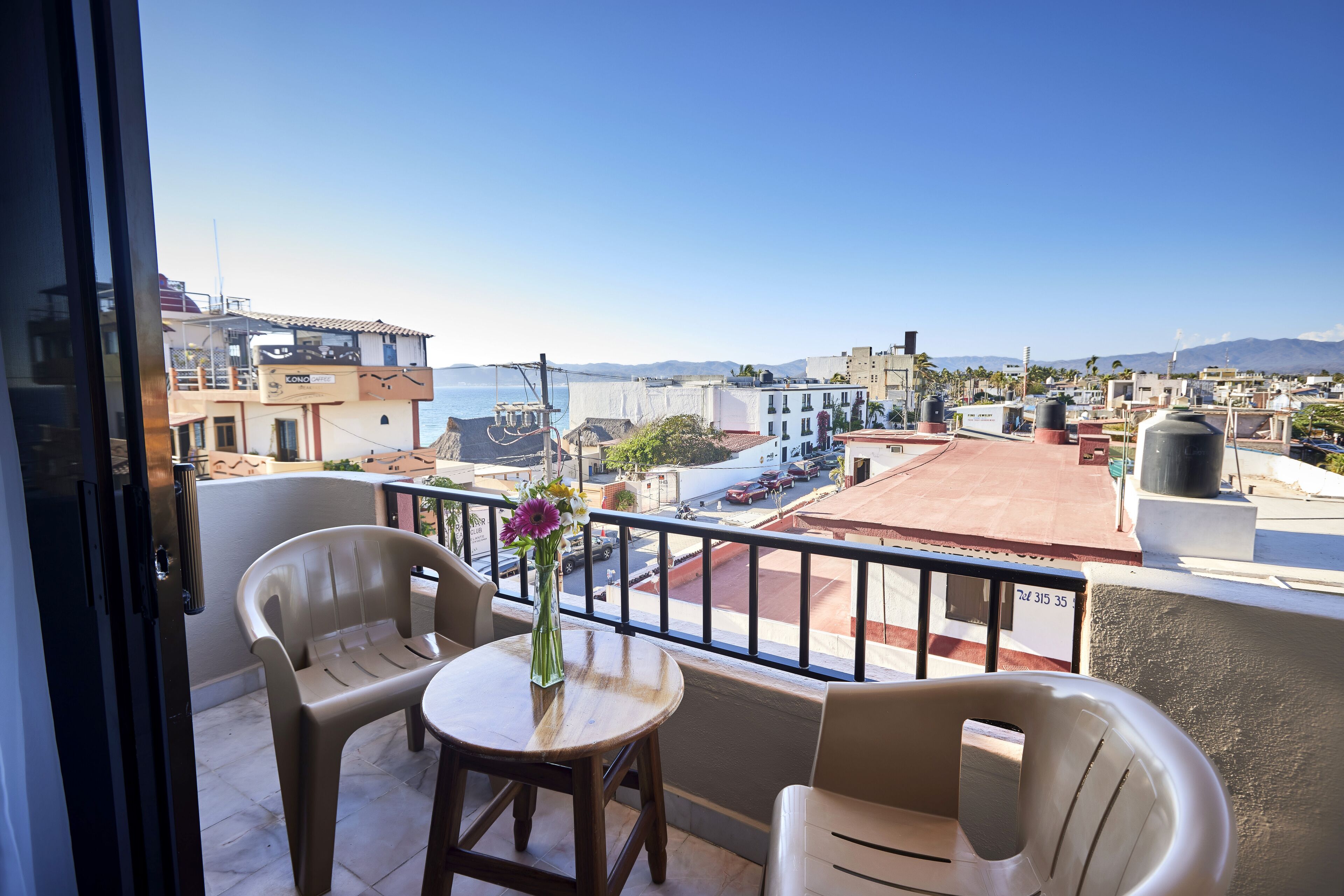 A balcony with a view of the city and sea, featuring plastic chairs, a small table, and a vase with flowers.
