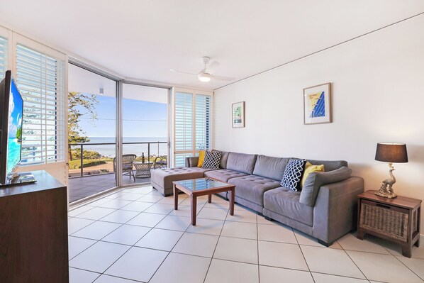 A spacious living room with a grey sofa, a coffee table, and a TV on a stand. There are large windows offering a view of the sea. - C Bargara Resort (Bargara)
