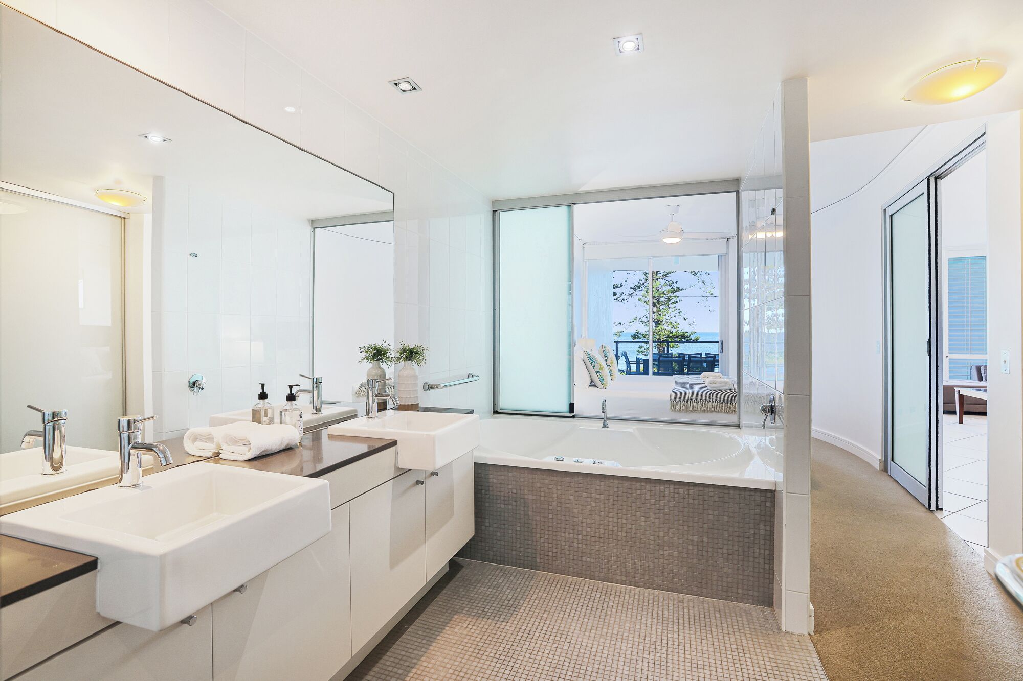 A modern bathroom with a large mirror, two sinks, a bathtub, and a view of the outdoors.