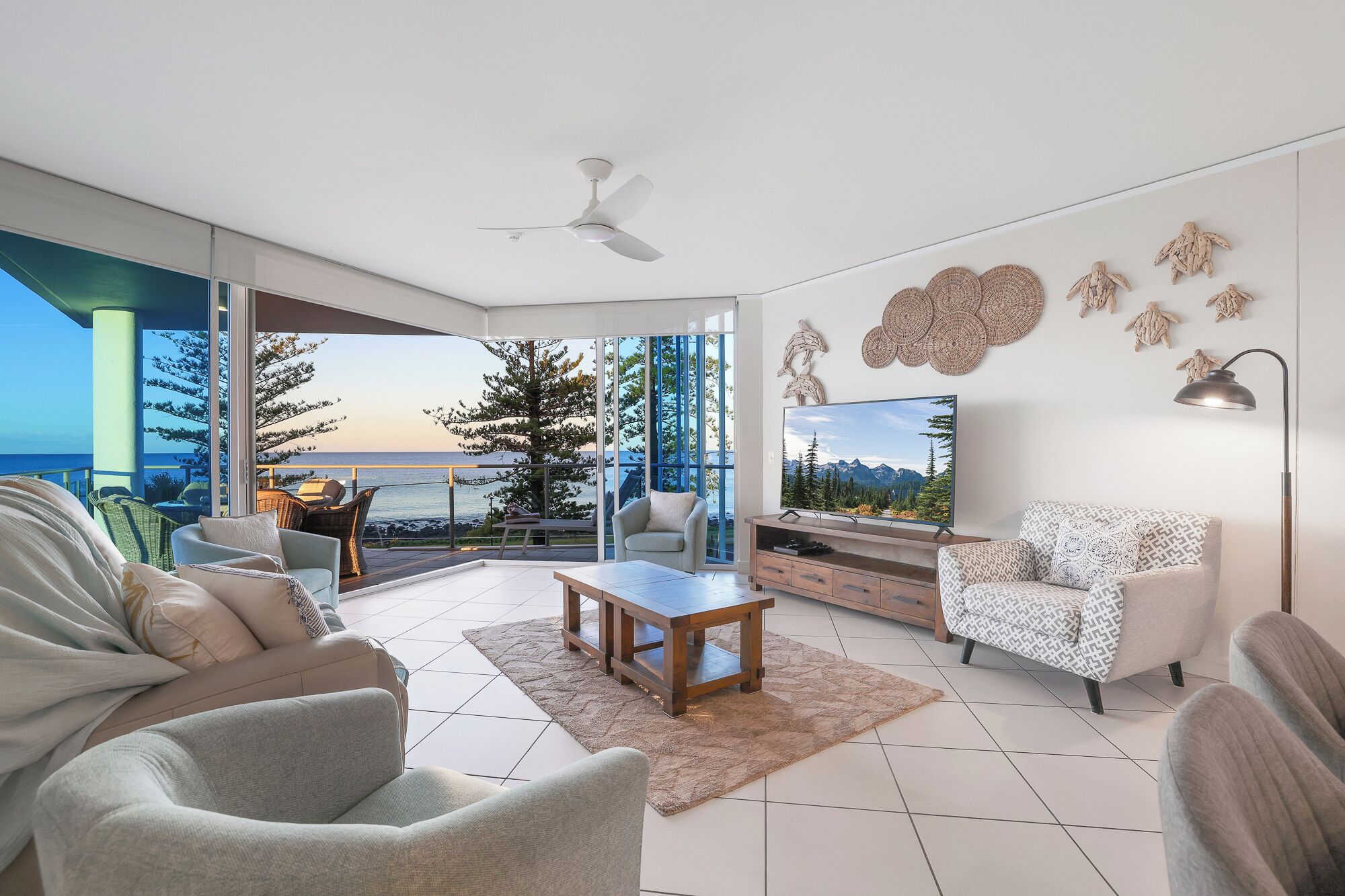 A modern living room with a large TV, comfortable seating, a ceiling fan, and a view of the ocean.