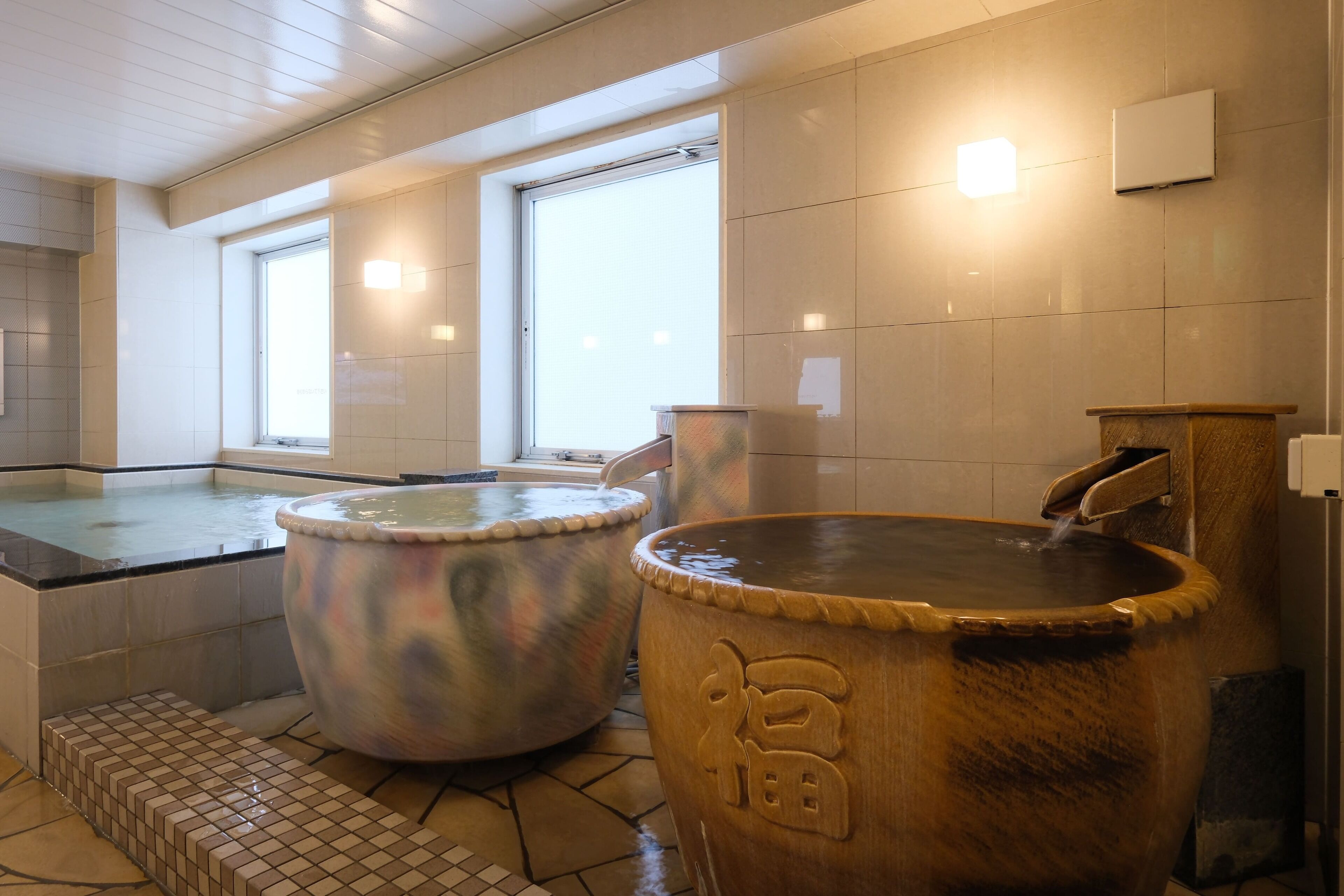 A traditional Japanese onsen with two large hot tubs, a tiled floor, and a window providing natural light.
