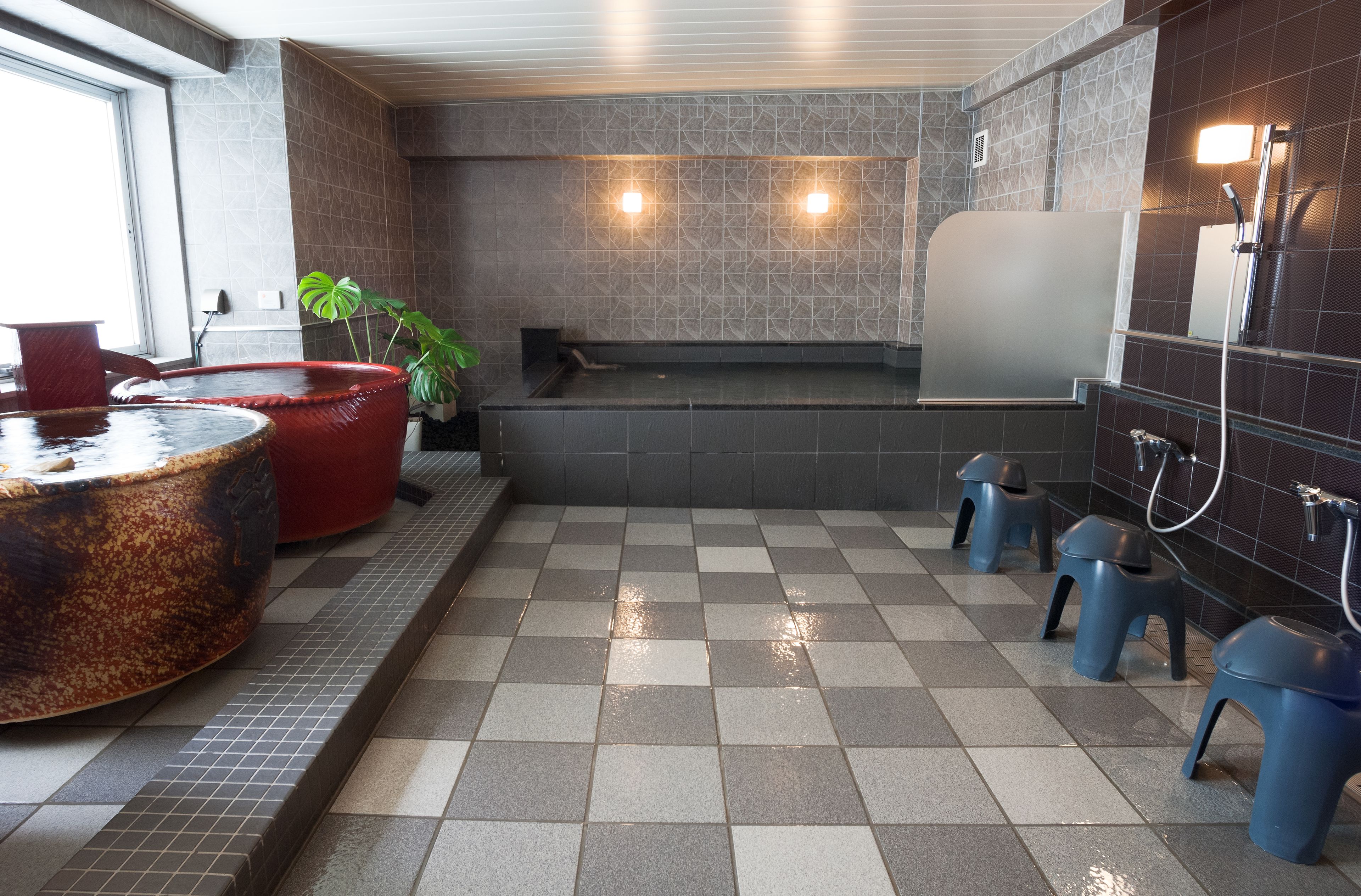 Eine öffentliche Toilette mit Schachbrettboden, roten und blauen Hockern, einem großen Waschbecken und einem wandmontierten Spiegel.