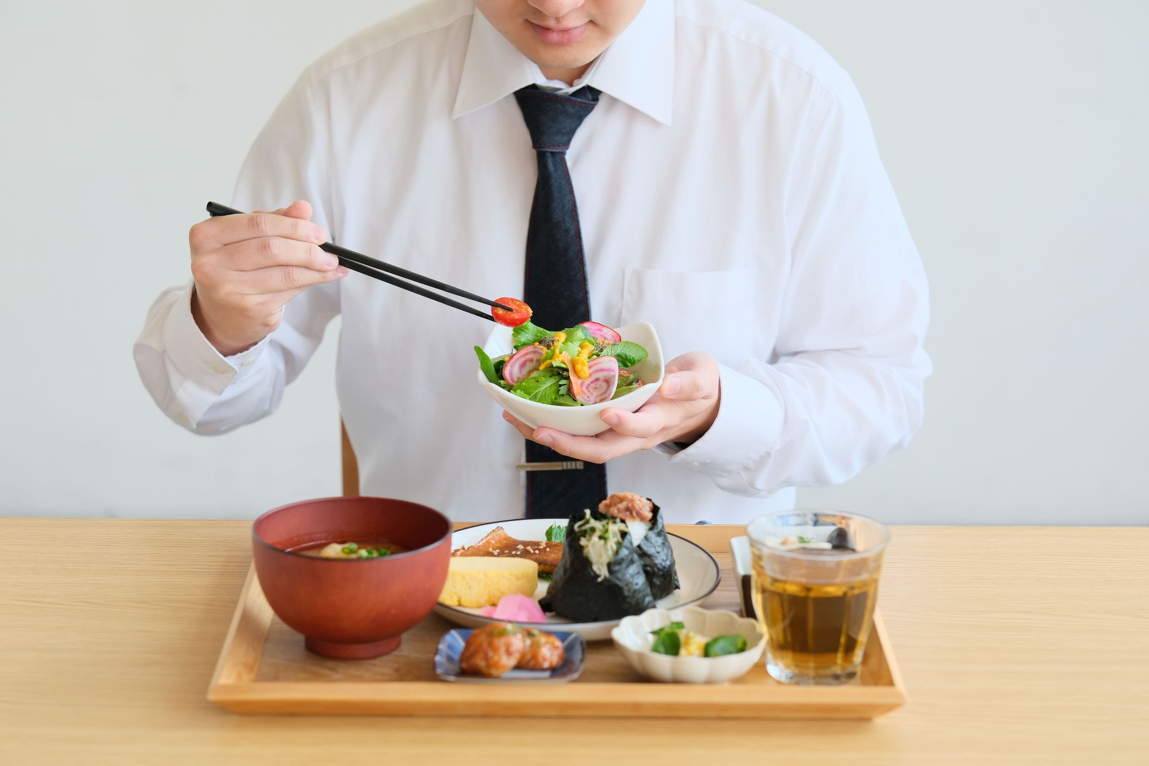A person holding a bowl of salad with chopsticks, a red bowl of soup, a plate with various dishes, and a glass of tea on a tray.