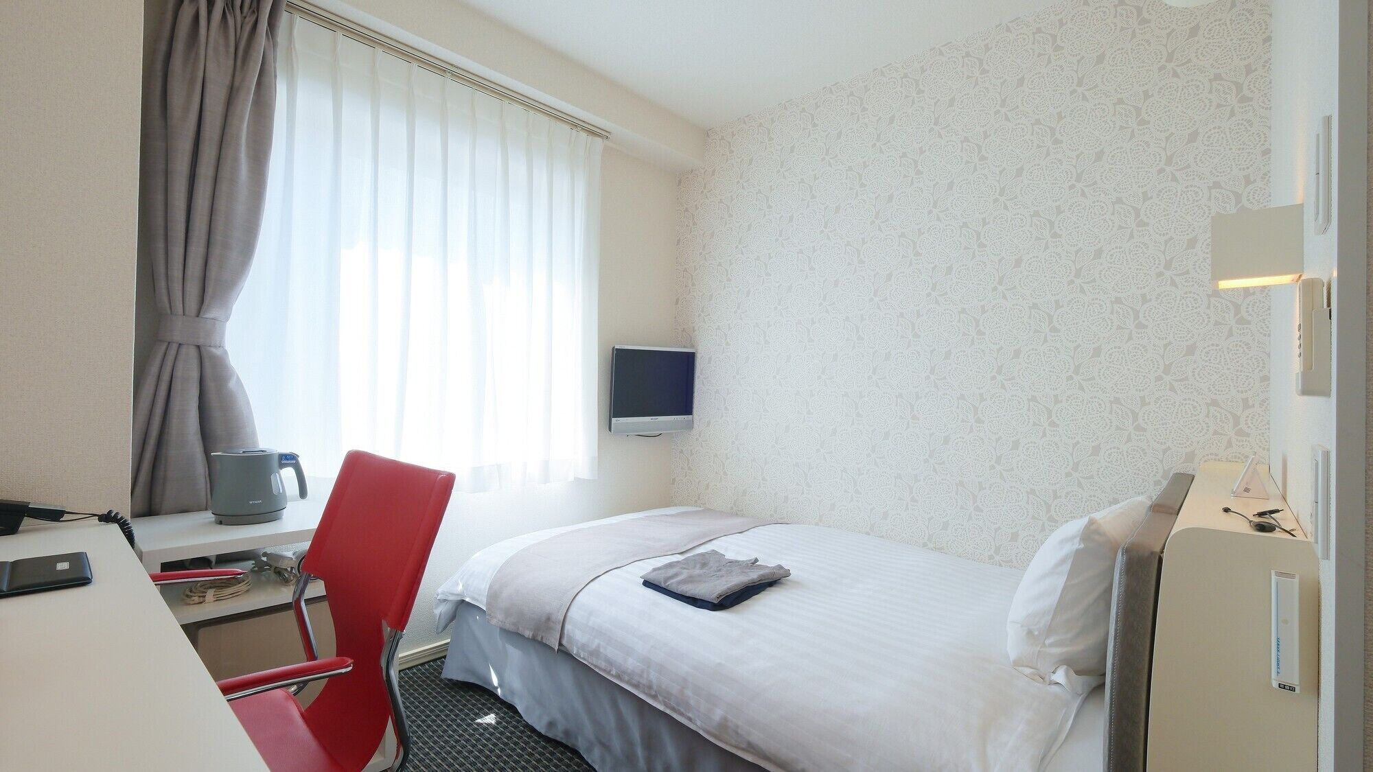 Ein Hotelzimmer mit Bett, Schreibtisch mit Computer, Stuhl, Fernseher und Fenster mit Vorhängen.