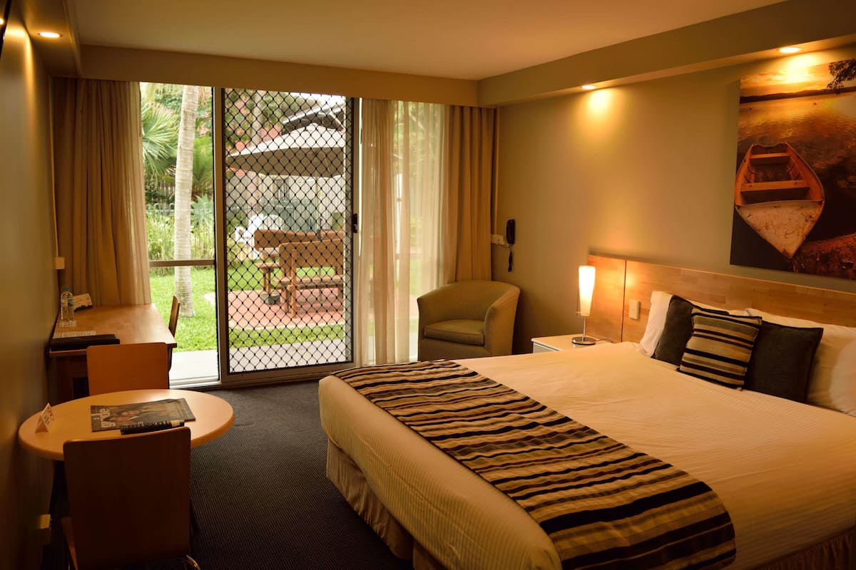A hotel room with a large bed, a desk with a chair, a small table, a telephone, and a view of the outdoors through a sliding glass door.