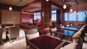 Un restaurant au décor moderne, avec des tables et des chaises en bois, des luminaires suspendus et de grandes fenêtres laissant entrer la lumière naturelle.