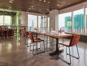 Un comedor moderno con una larga mesa dispuesta para seis personas, rodeada de sillas de cuero, grandes ventanales con vista a la ciudad y un techo alto con iluminación empotrada.